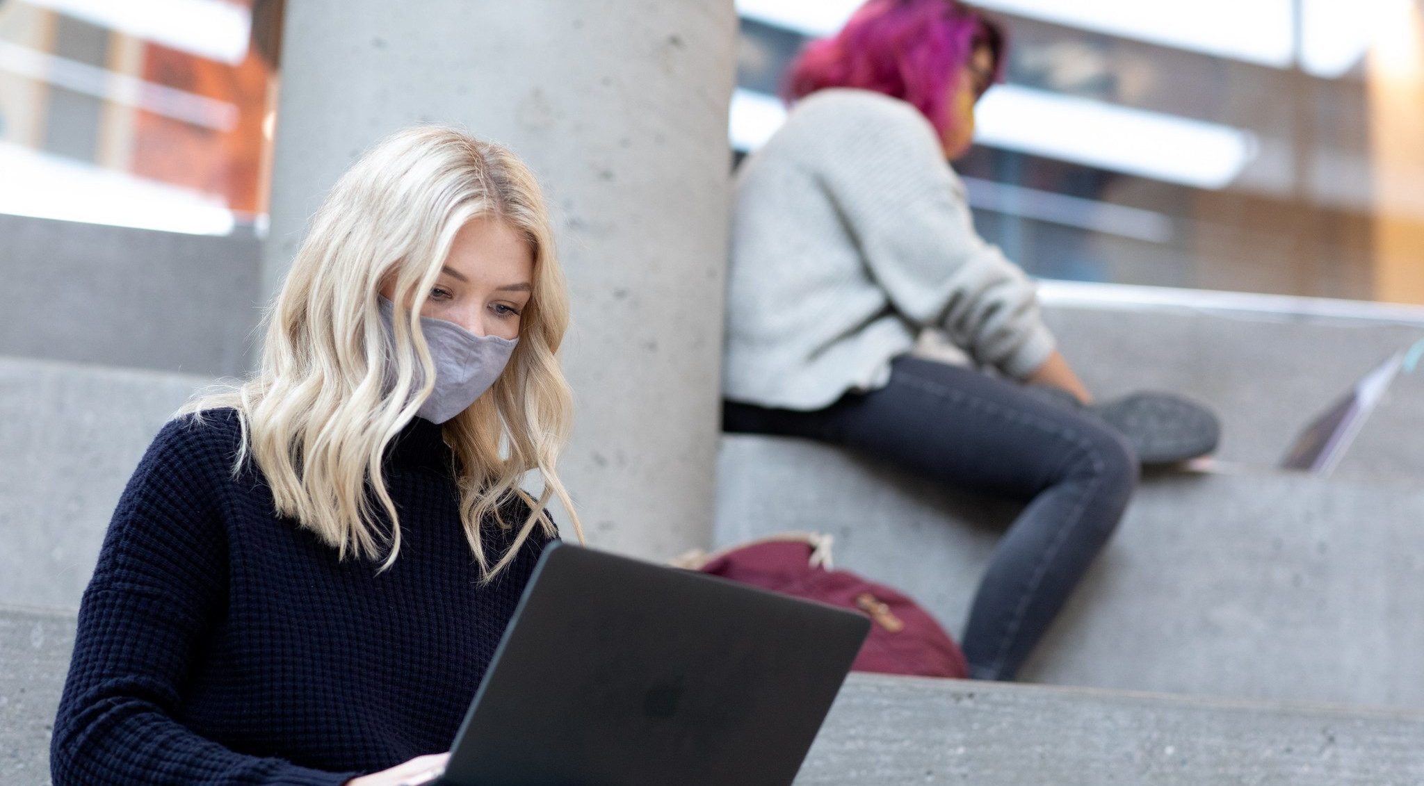 Two students sit on concrete steps inside the Fipke building. In the forebround, a student with blonde hair and a grey cloth mask looks down at their laptop. In the background, a student with pink hair does the same.