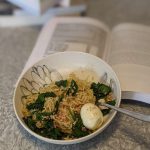 A bowl of instant ramen holding open a textbook with spinach and a boiled egg added to it