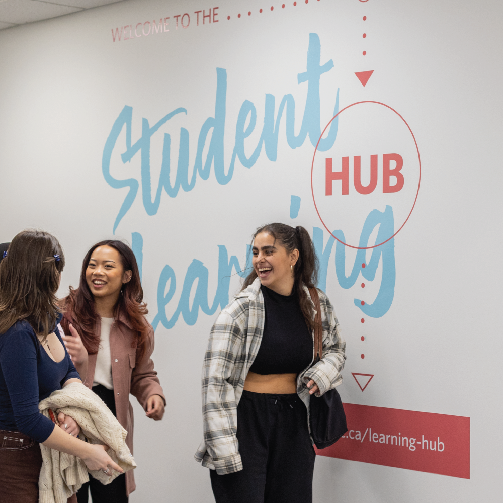 three UBCO students walking into the Student Learning Hub on campus. They are looking at each other and laughing with the Student Learning Hub logo on the wall behind them